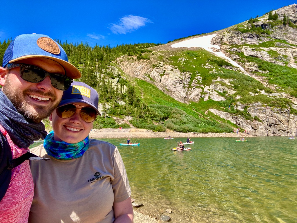 Husband and wife selfie with lake and paddle boarders in background.